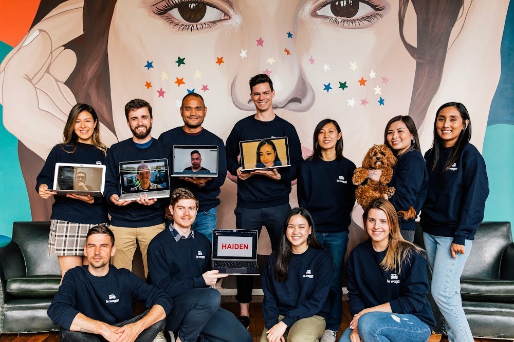 A group of diverse young adults, dressed in matching dark sweatshirts, poses joyfully in front of a vibrant mural featuring a large face and colorful stars, some holding open laptops displaying video call participants, with one person holding a small brown dog; team spirit and remote collaboration are highlighted.