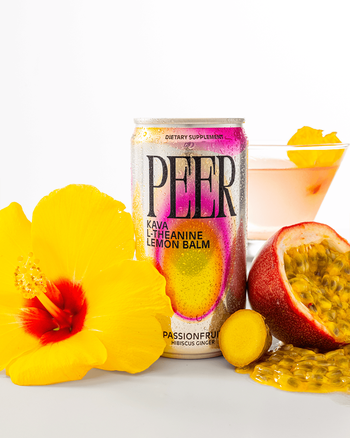 A can of Peer Kava L-Theanine Lemon Balm is styled with yellow hibiscus flowers, a passion fruit, and a pink cocktail glass on a clean white background.