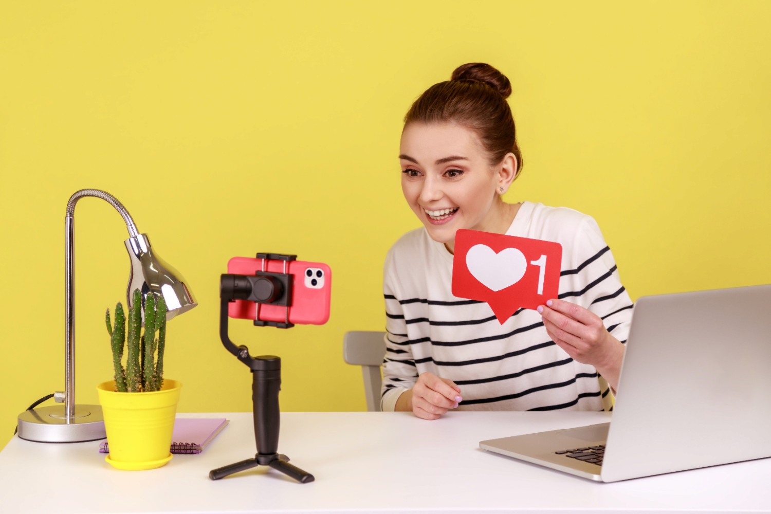 A woman holds a heart-shaped object while sitting in front of a laptop, symbolizing love and technology.