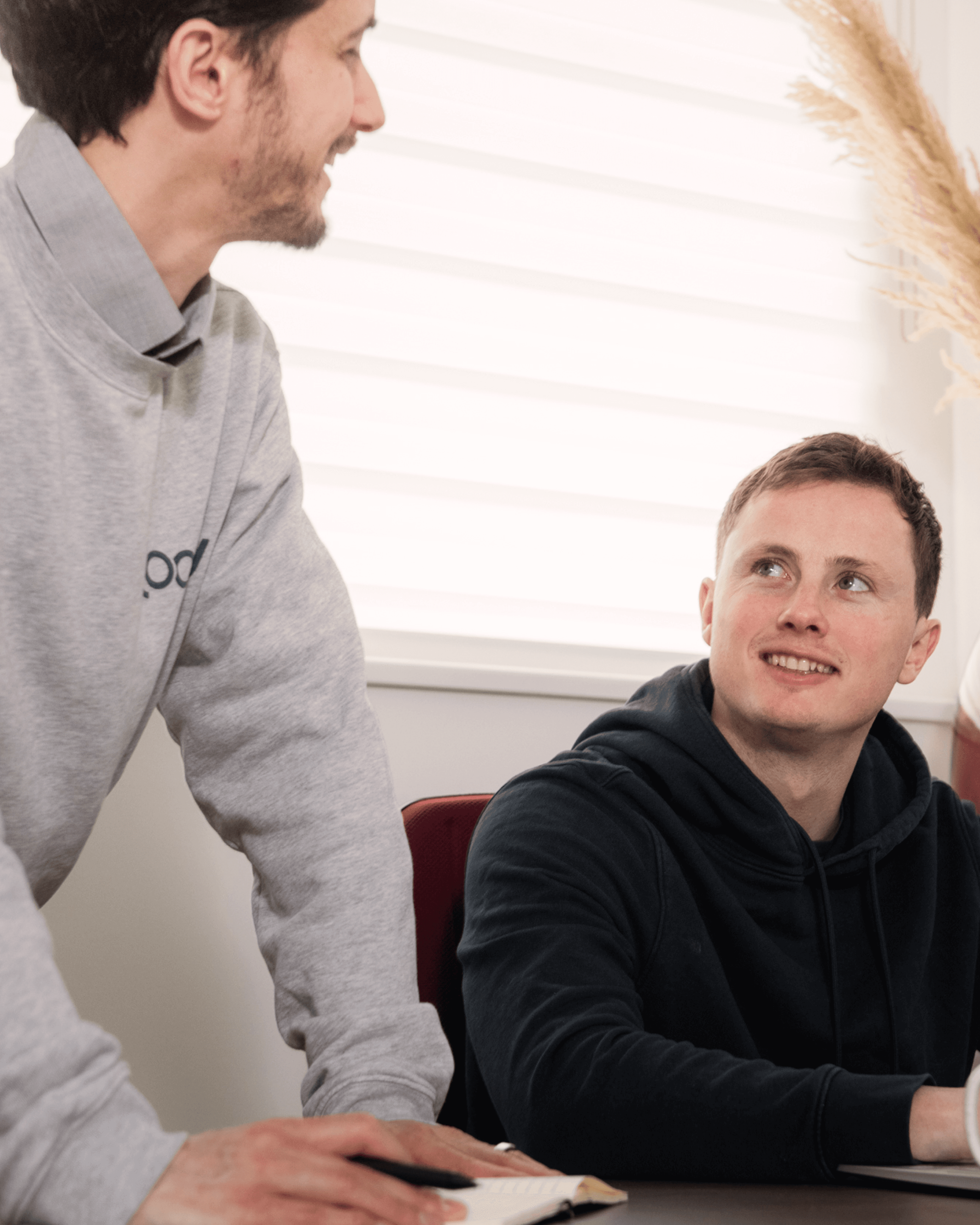 Two men engage in conversation, one standing and leaning on a table, the other seated and listening attentively.