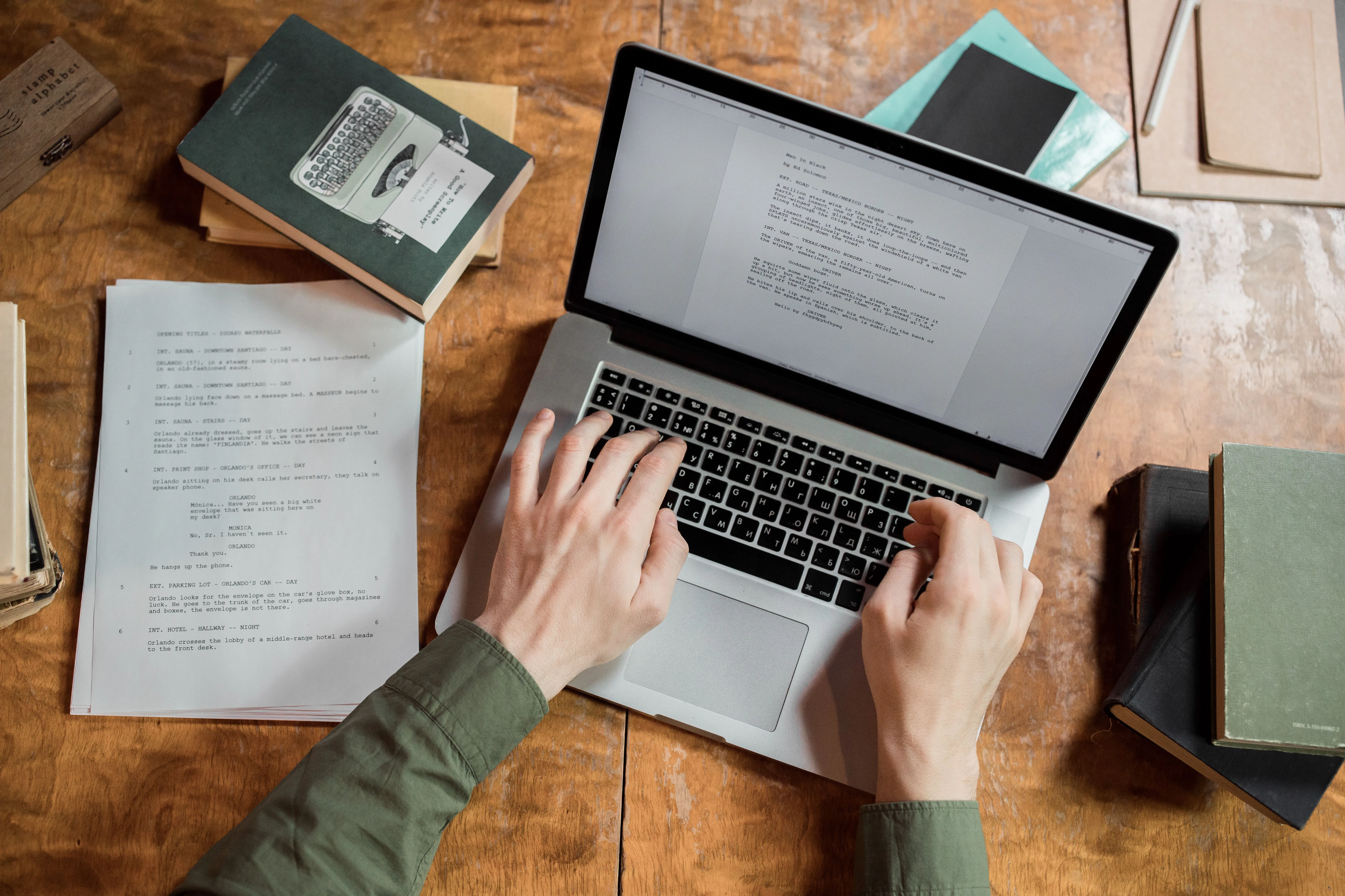 A person wearing a green shirt types on a laptop surrounded by stacks of books and notebooks on a wooden table, emphasizing a focused study or work environment.