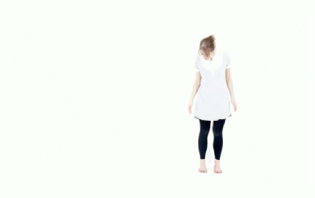 A woman with light brown hair pulled back in a loose bun stands barefoot against a plain white background. She wears a white short-sleeved top and dark-colored leggings or pants. Her arms are relaxed at her sides, and her head is turned slightly to the right. The image focuses on her upper body and back.