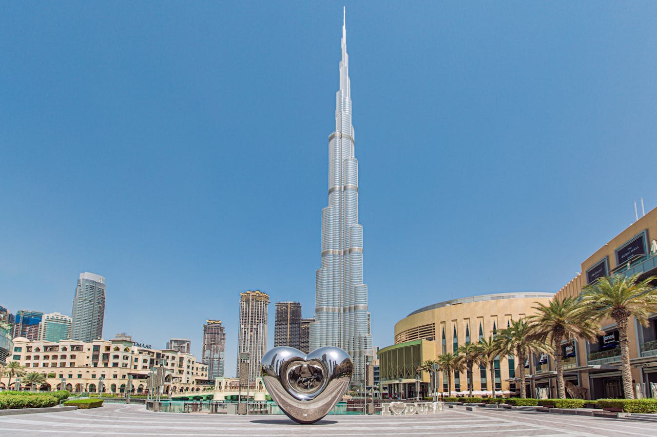 The Burj Khalifa over Downtown Dubai on a bright sunny day, with palm trees, buildings, and a heart-shaped structure in the foreground.&nbsp;