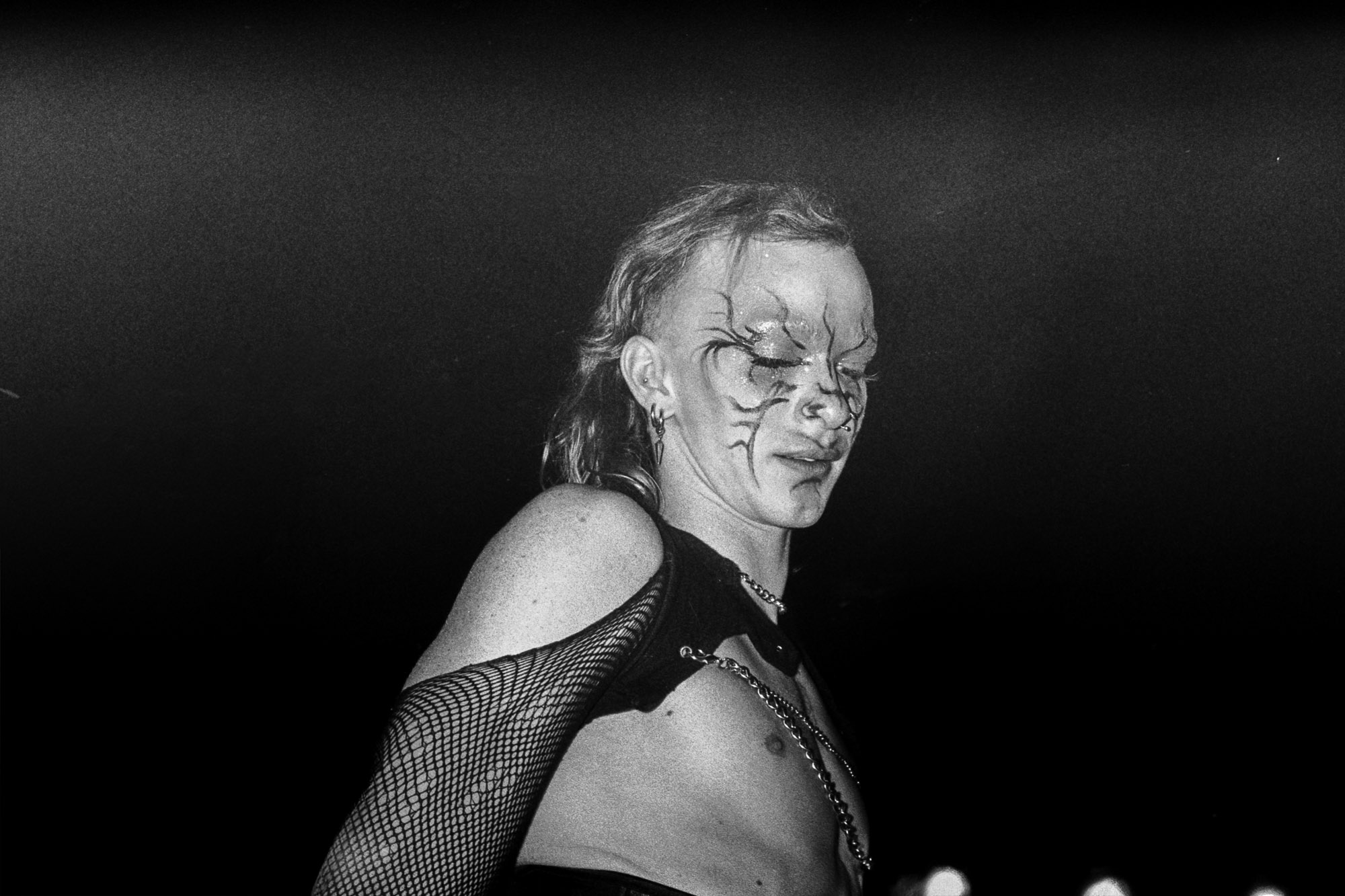 A person with dramatic face paint and a mesh sleeve stands under dim lighting, exuding a gothic and theatrical presence.