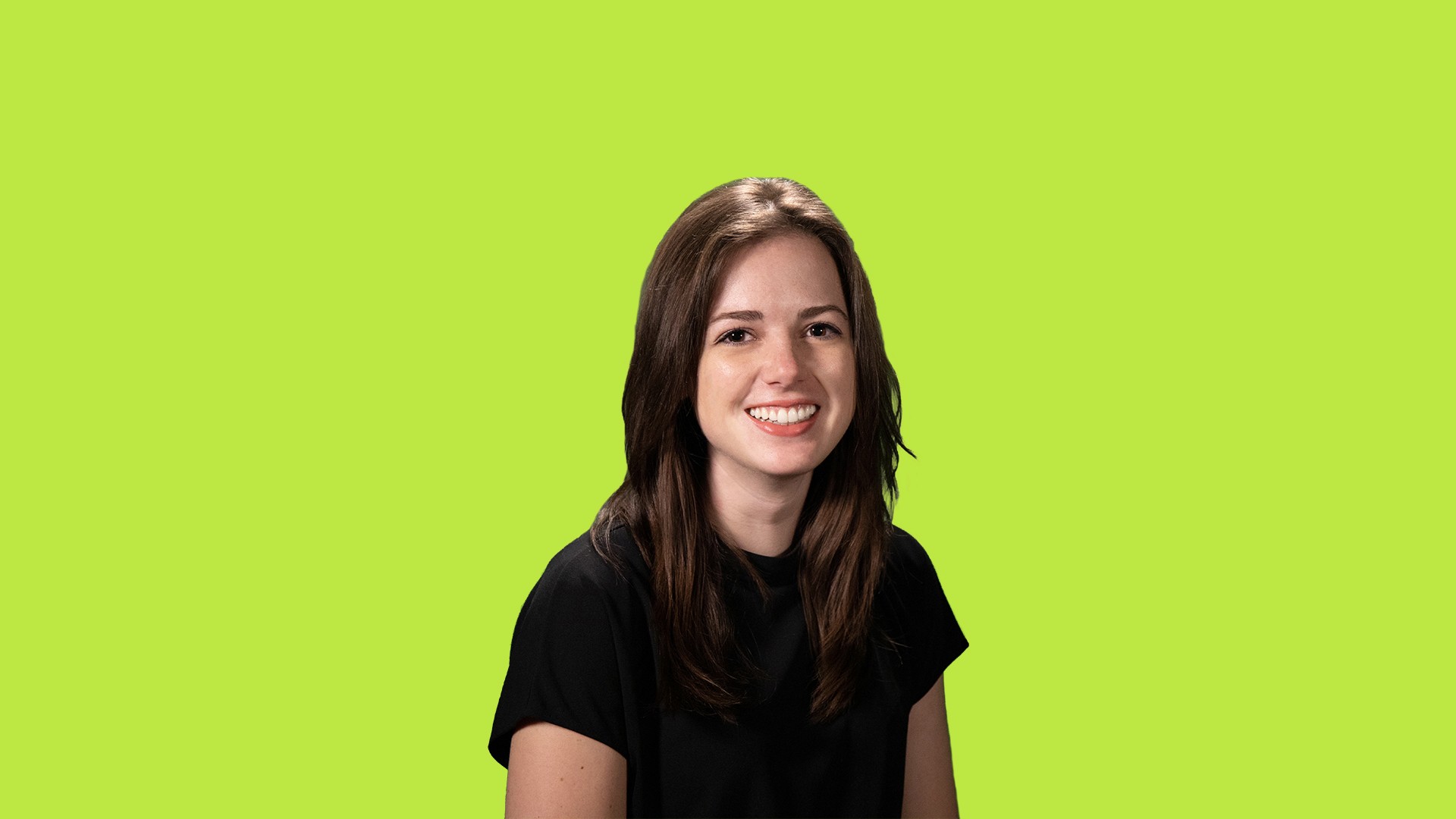 A woman with long brown hair wears a black short-sleeved t-shirt against a bright green background. She is smiling and looking directly at the camera.