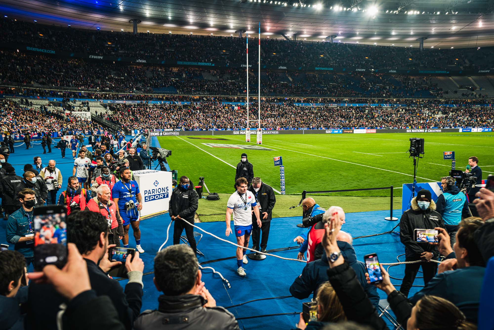 Antoine Dupont en tee-shirt blanc, médaille au cou, quitte la pelouse escorté de stadiers, entouré de photographes et de spectateurs qui le filment