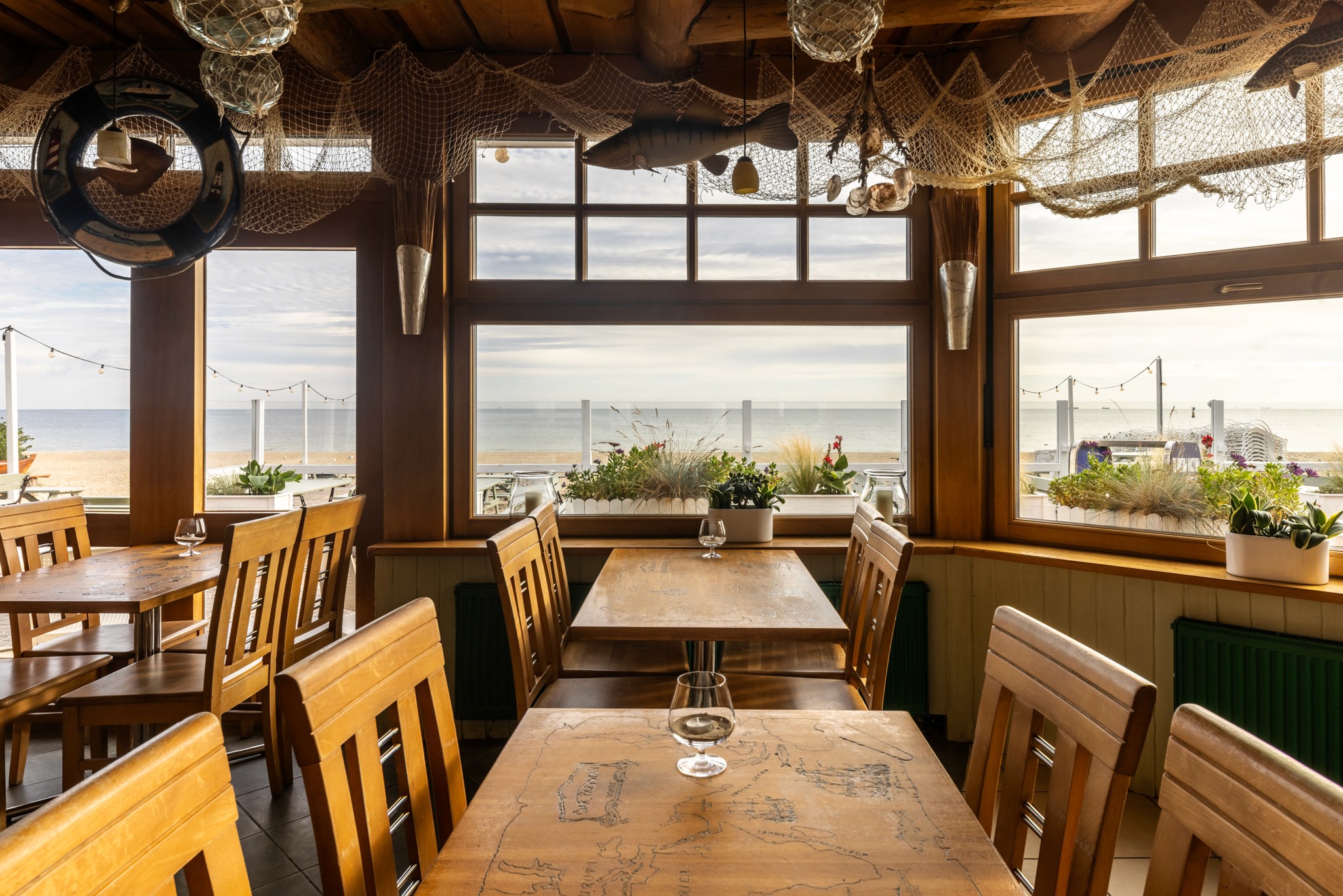 Restauracja nadmorska z drewnianymi stołami i krzesłami, marynistycznym wystrojem oraz dużymi oknami z widokiem na plażę i morze.