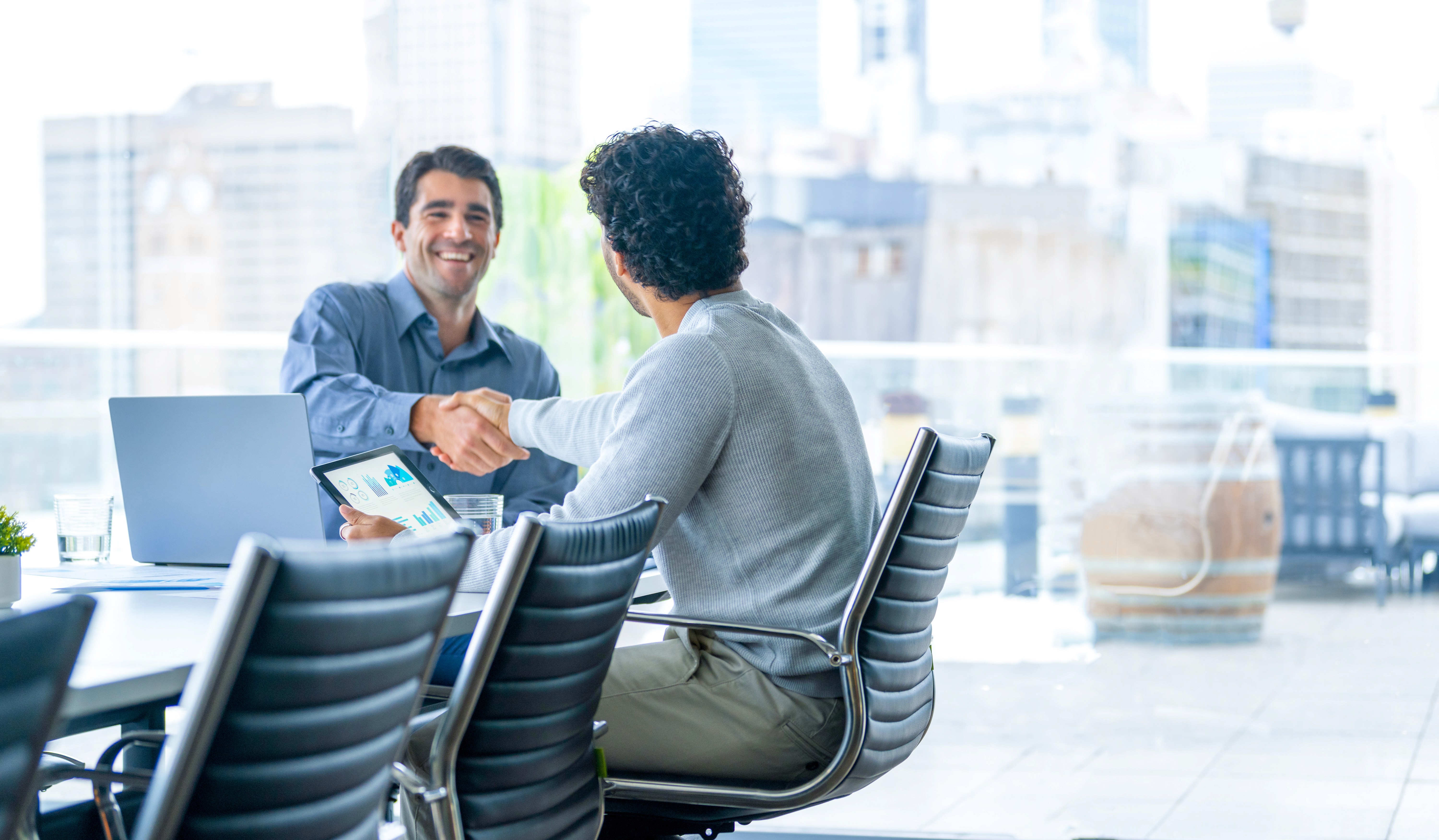 Two businessmen shaking hands at a boardroom table with analytics on a tablet