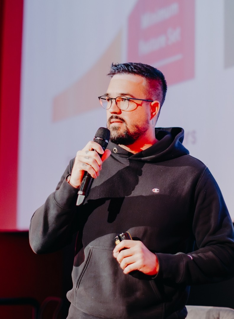 Man in a sweater smiling with arms crossed while standing by a wooden wall.