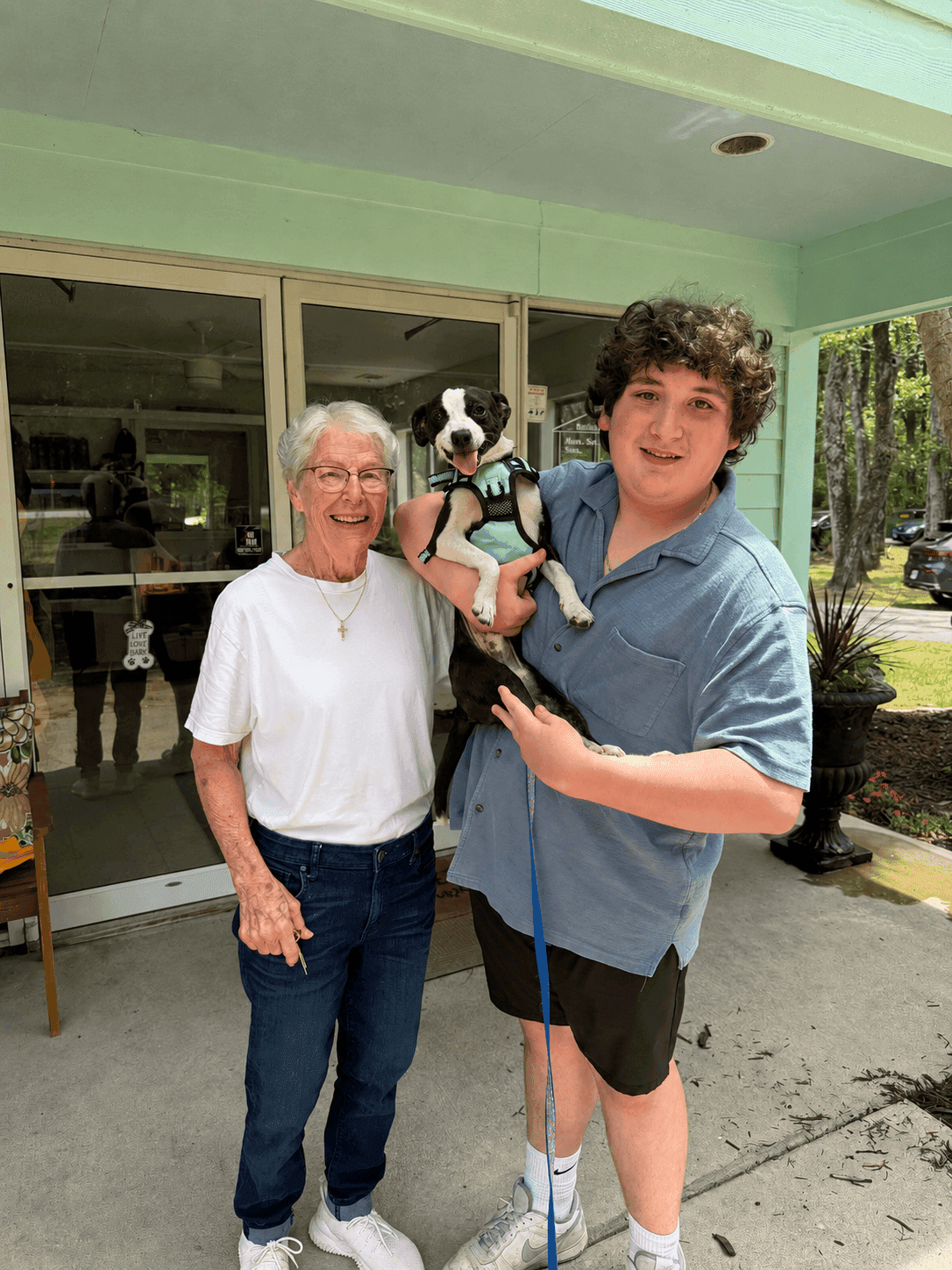 A young man and an elderly man pose together outdoors, smiling, with a small dog in the young man's arms.