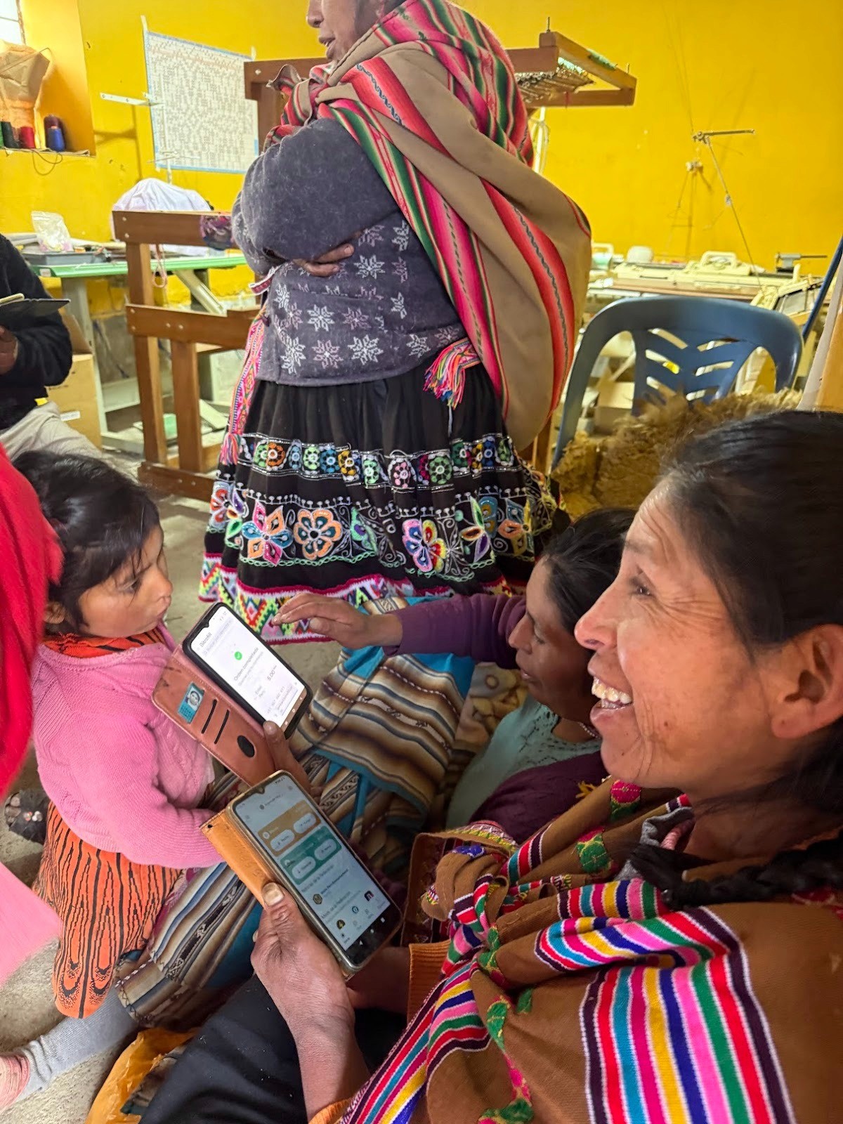 An Andean woman in traditional dress smiles as she explores the Fedi app on her phone, while a young child and other community members in colourful traditional clothing look on and engage with their own devices nearby.