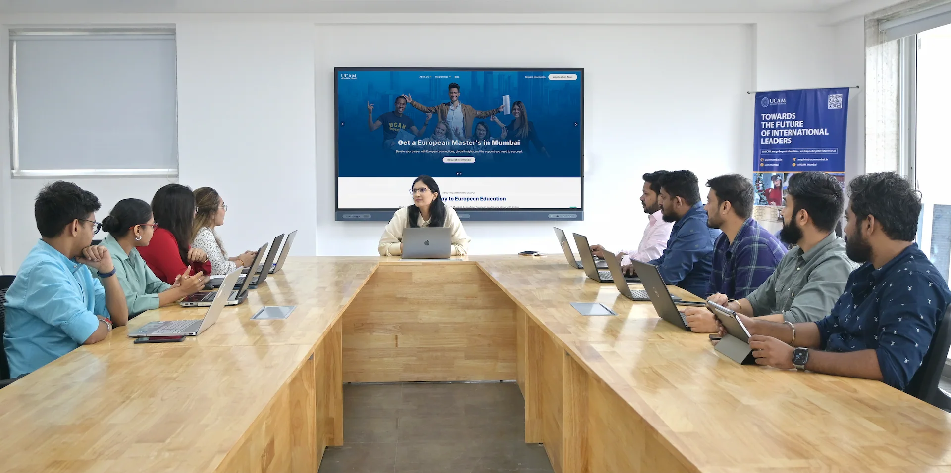A diverse group of students is seated in a modern classroom at a large wooden table, all with laptops, attentively listening to a lecturer at the head, with a screen in the background displaying information about European master's programs, emphasizing the UCAM Mumbai campus.
