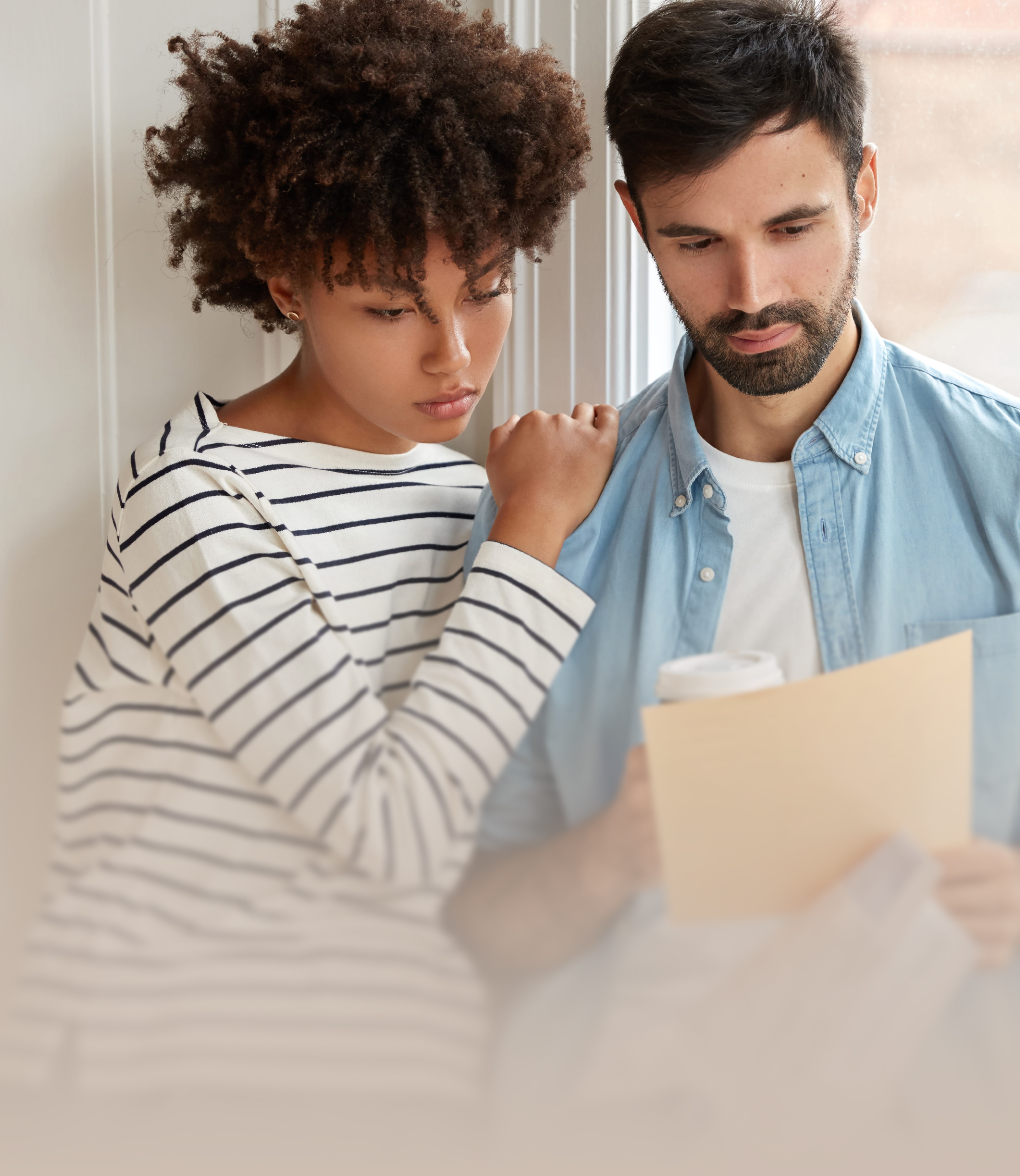 A man and woman stand close together by a window, intently reviewing a document, with the woman in a striped shirt and the man holding a coffee cup, creating a collaborative and focused atmosphere.