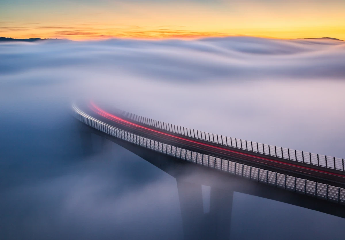 Aerial view of the Črni Kal viaduct in Slovenia at sunset, with its tall concrete pillars rising above a thick sea of fog in the valley against a vibrant orange and blue twilight sky.