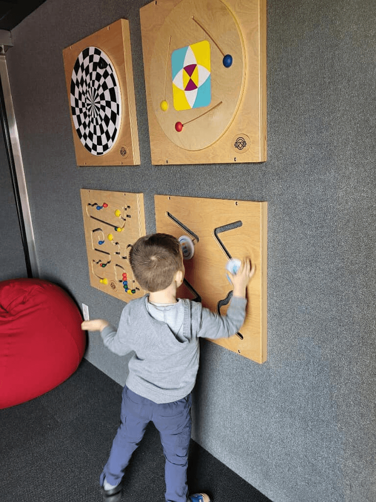A young boy seen from inside the Sensory Activation Vehicle, reaching up to guide pieces along an interactive wooden wall maze. Three other colorful sensory wall boards are mounted nearby on the gray felt wall next to a red beanbag chair.