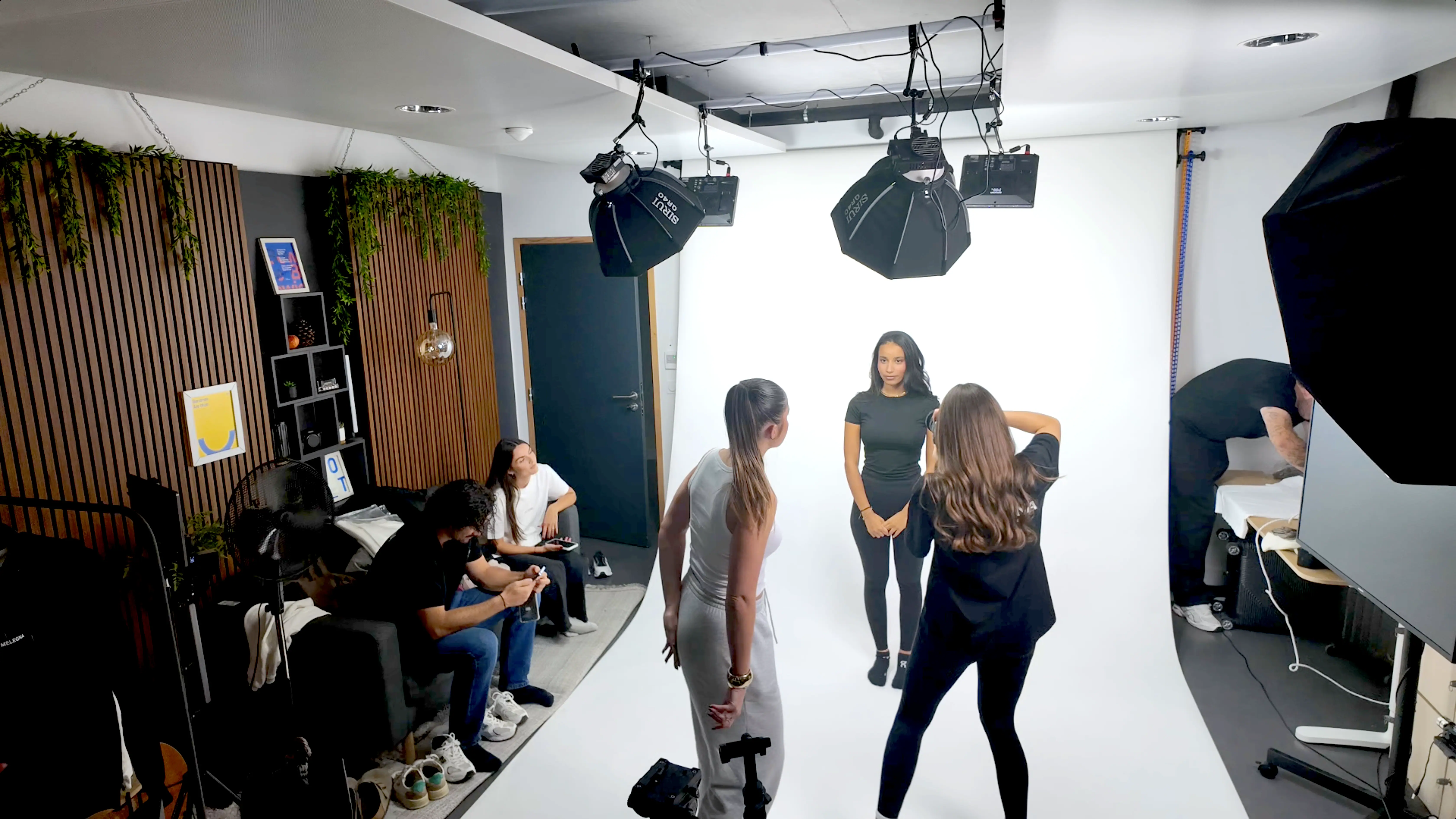In a professional photography studio, a subject poses against a white backdrop while two individuals converse near the setup, surrounded by equipment and studio lights.