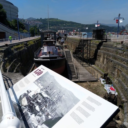 An information board overlooks a dry dock containing two boats, with a backdrop of buildings and hills under a clear sky.
