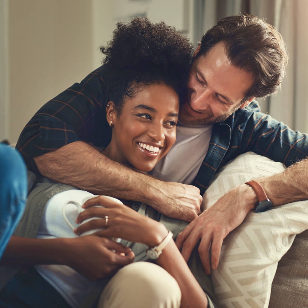 A smiling couple embraces on a couch. She holds a mug, wears a ring. He's in plaid.