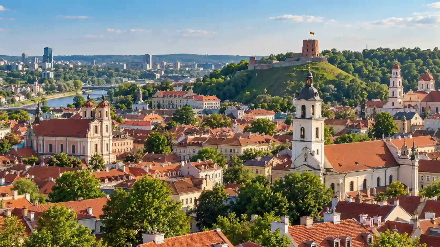 Panoramic daytime view of Vilnius Old Town in Lithuania with red rooftops, historic church towers, green spaces and Gediminas Castle Tower on the hill.