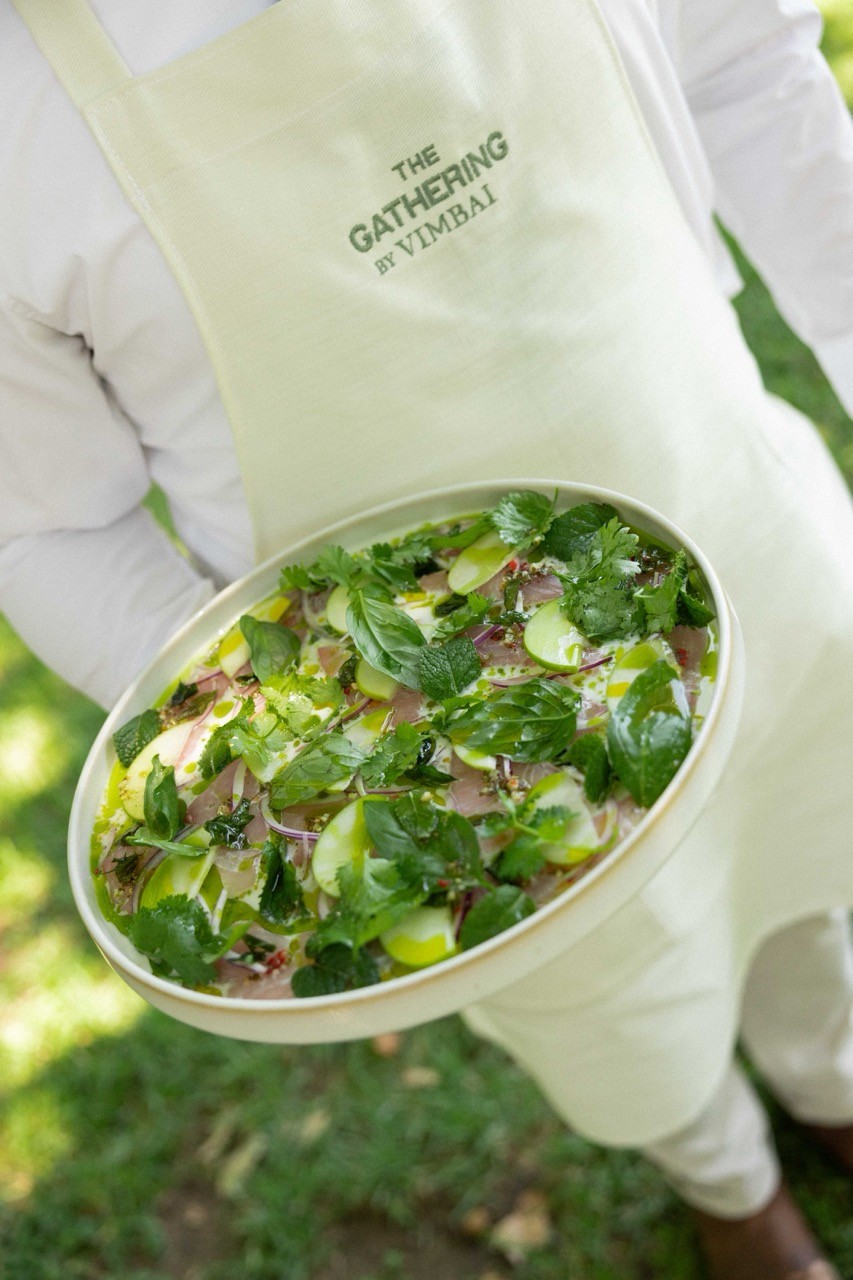 Close-up of server's hand holding plate labeled "THE GATHERING AT VIMBAI" featuring fresh herb salad with cilantro, greens, and delicate plating on white dish