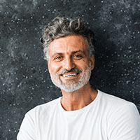 A smiling man with curly hair and a beard stands against a dark speckled background, wearing a white t-shirt. The mood is friendly and relaxed.