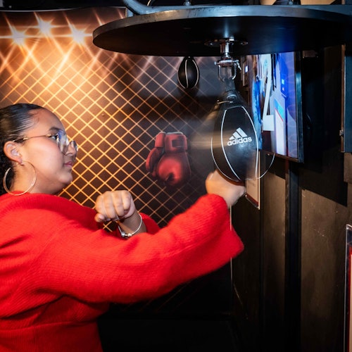 En person i en rød trøje slår på en boksebold i et sportstræningsområde med boksehandsker hængende i baggrunden.