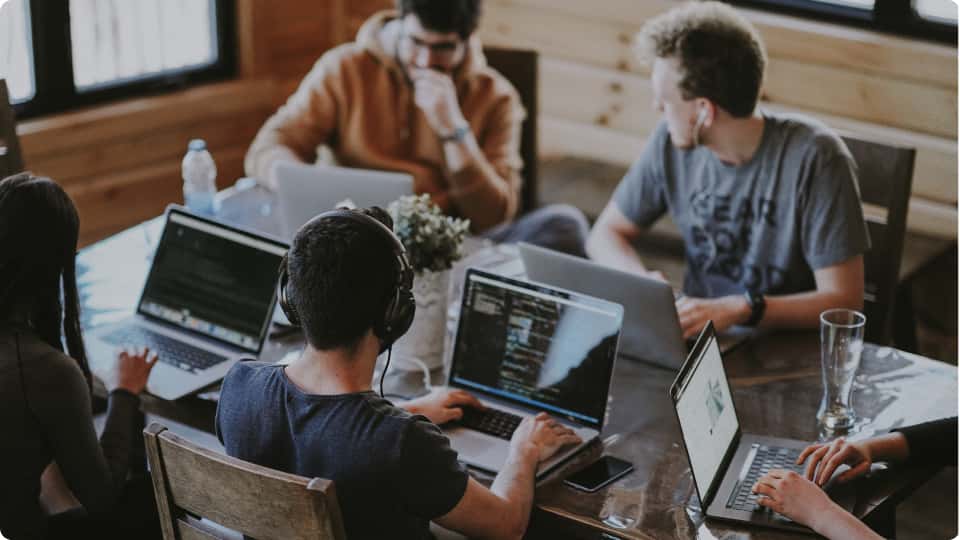 Diverse group of young professionals seated at a wooden table, engaged in collaborative work on their laptops.