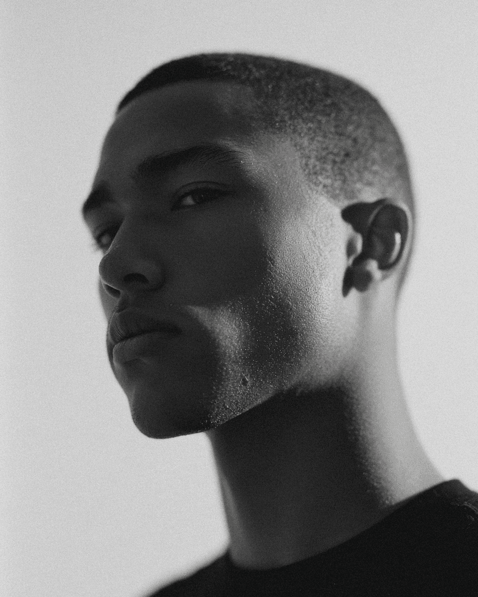 Black and white editorial portrait of a man in side profile with dramatic shadow, short hair, soft film grain, and minimalist studio background