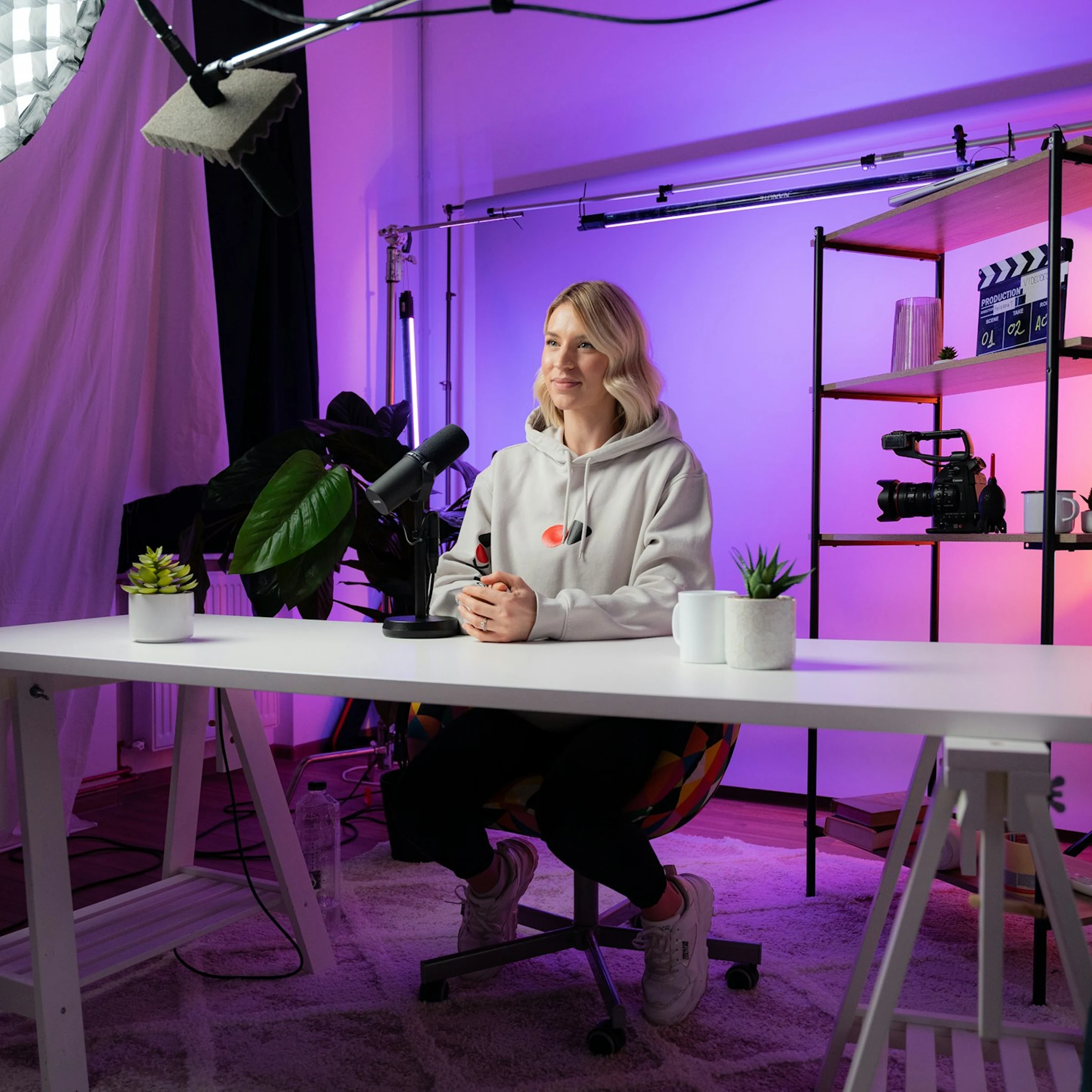 Woman in a white hoodie seated at a desk in a small purple-lit studio