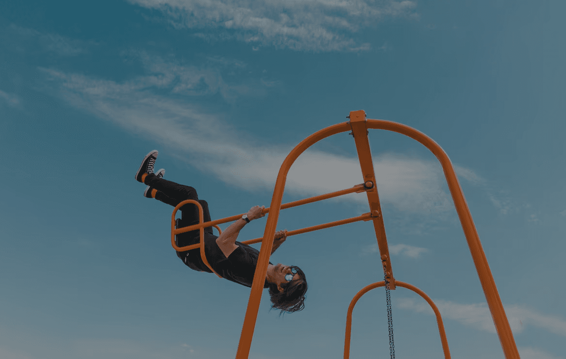 Person hanging upside down from orange playground bars against a bright blue sky.