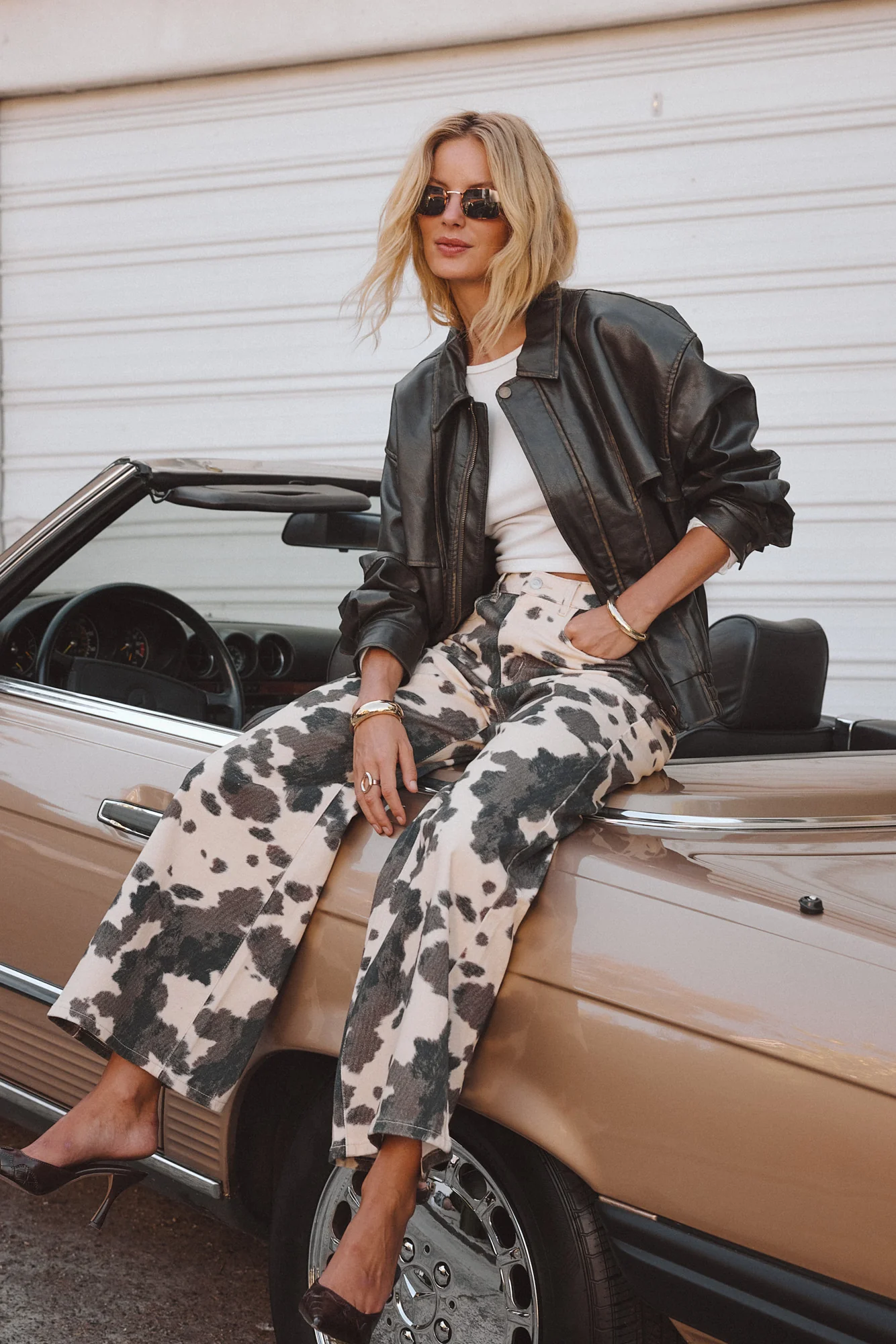 Person wearing sunglasses and a bomber jacket sits on a vintage car, styled with patterned pants.