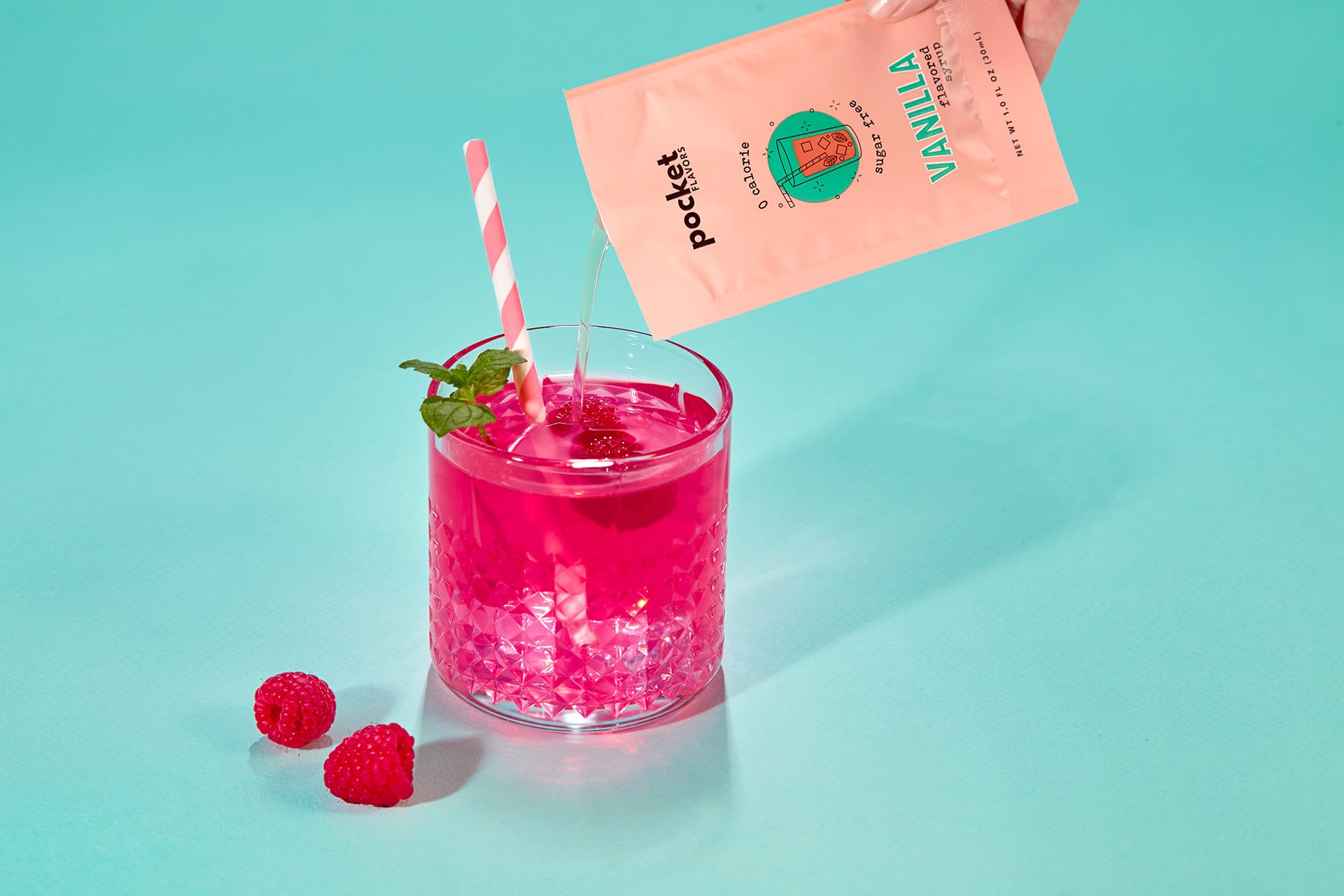 Ręka trzyma i wlewa paczkę mieszanki waniliowej Pocket Flavors do szklanki z żywym różowym napojem malinowym, ozdobionym świeżą miętą i towarzyszącą pasiastą słomką, na tle turkusowego tła.