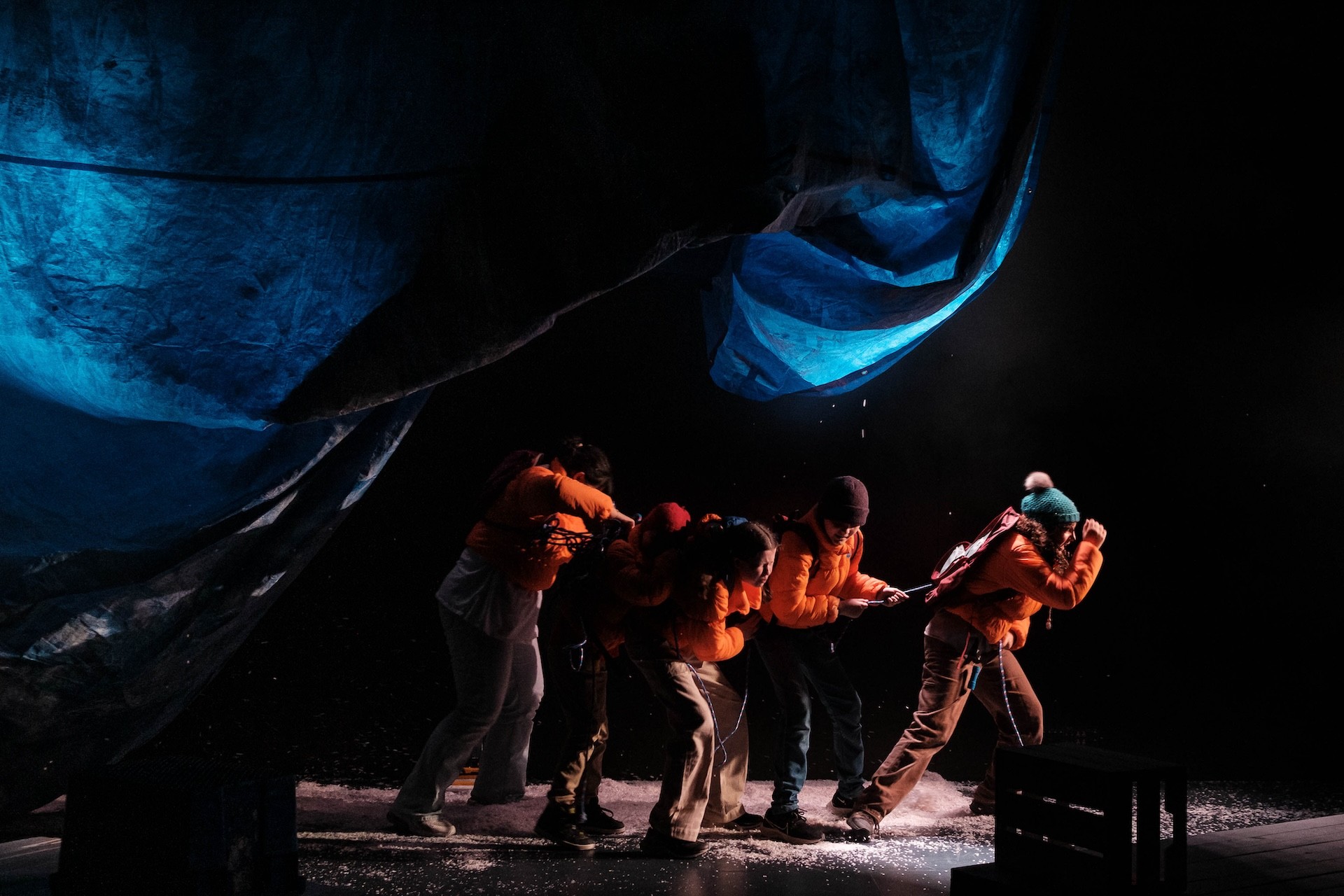 A line of performers in orange puffer jackets lean forward against a harsh wind effect, gripping a rope as artificial snow blows around them beneath a billowing blue tarp overhead.