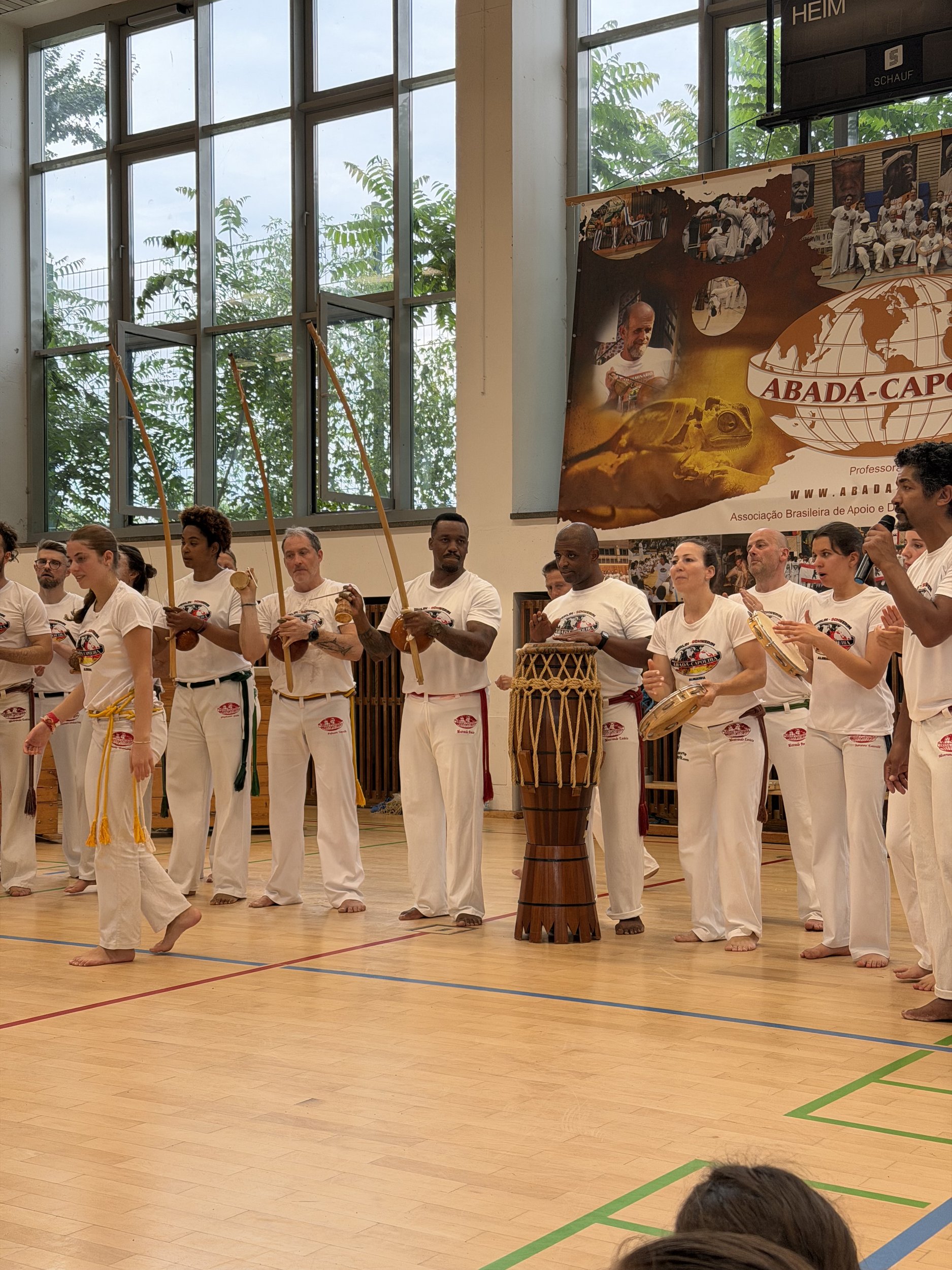Große Gruppe von Capoeiristas in weißen Uniformen spielt gemeinsam Berimbau, Atabaque und Pandeiro in einer Sporthalle vor einem ABADÁ-Capoeira-Banner.
