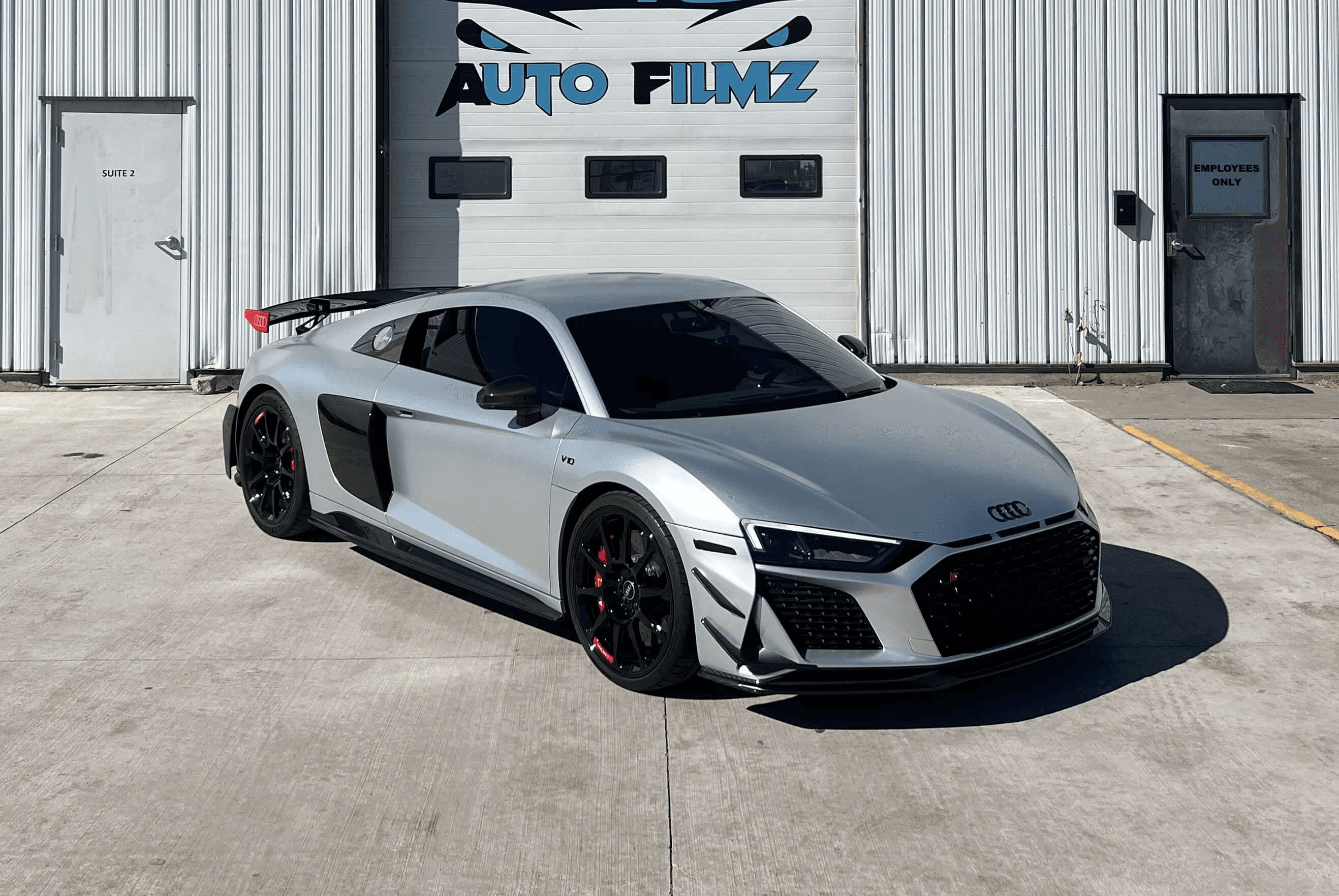 A sleek silver Audi R8 sports car is parked in front of a white industrial building with the sign "Custom Auto Filmz" displayed prominently above one of the garage doors.
