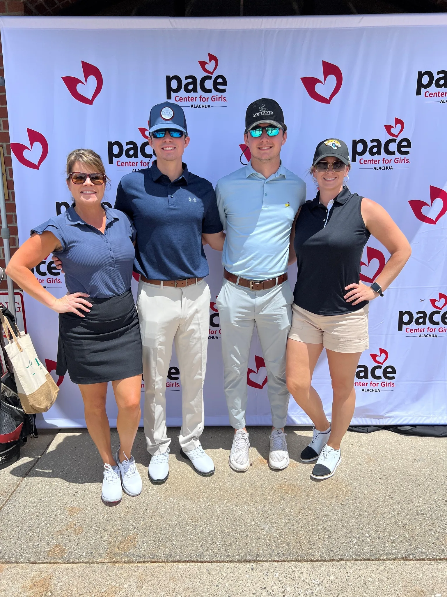 Four golfers in casual athletic wear stand together smiling in front of a PACE Center for Girls branded backdrop at what appears to be a charity golf event.