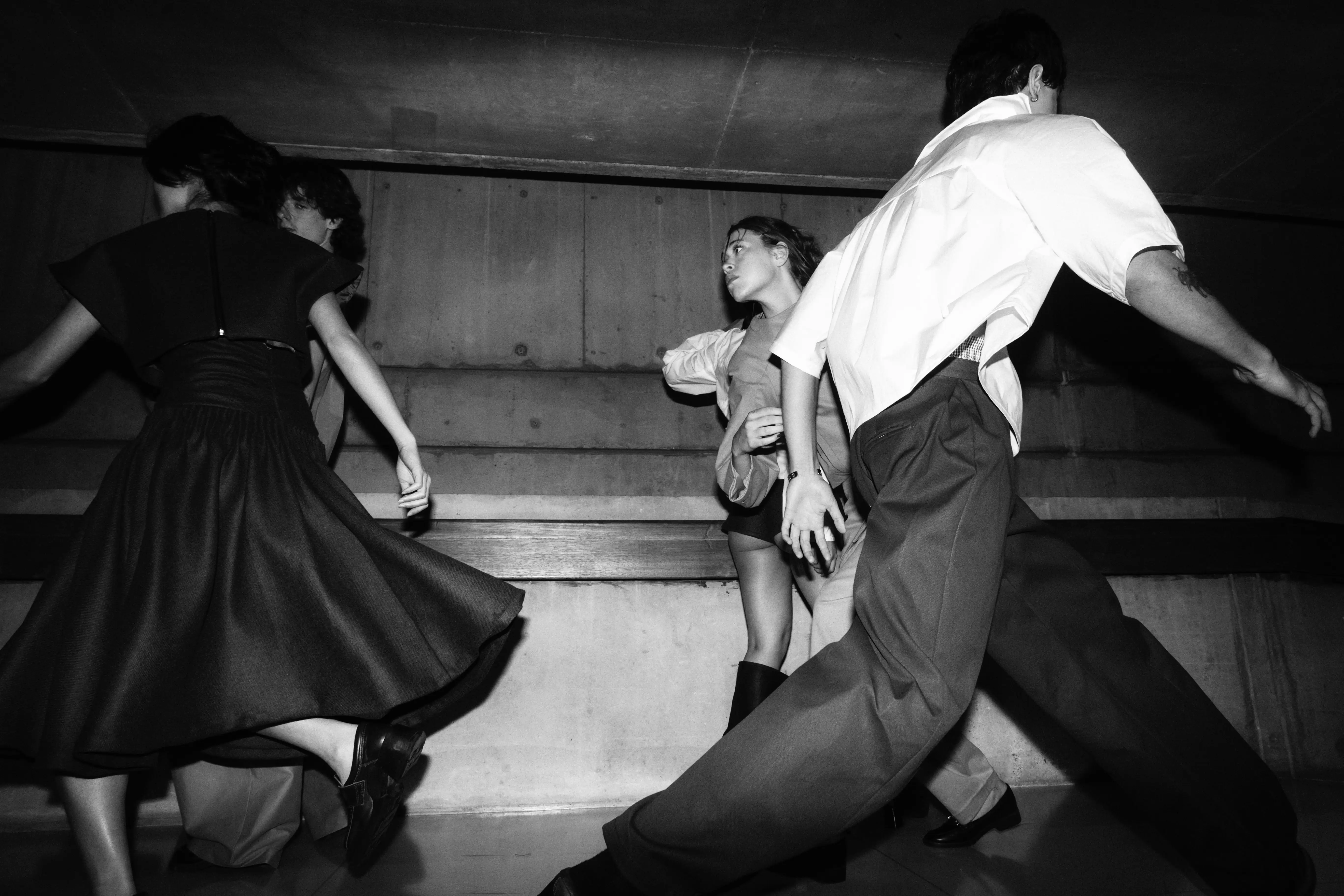 A black-and-white editorial shot of models in motion, wearing tailored skirts and button-down shirts while moving through a concrete, industrial space.