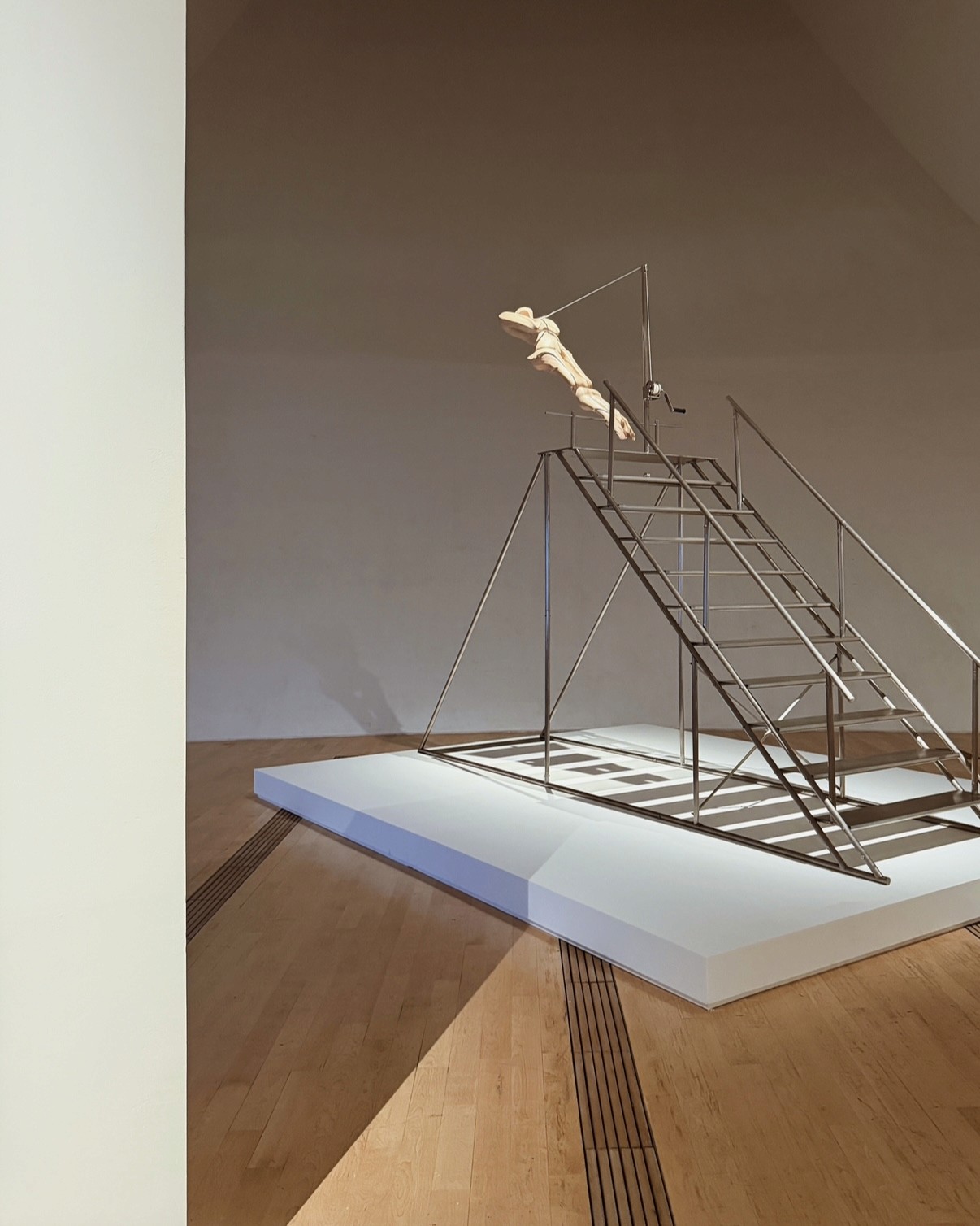 A contemporary art installation featuring a metallic structure resembling a staircase with a suspended wooden figure, set on a white pedestal in an art gallery.