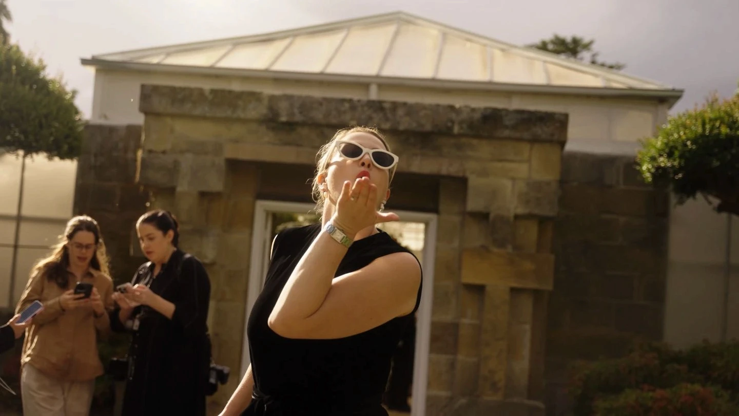 A person wearing sunglasses and a black outfit stands in front of a stone building, blowing a kiss towards the camera, while two other individuals engage with their phones in a sunny outdoor setting.