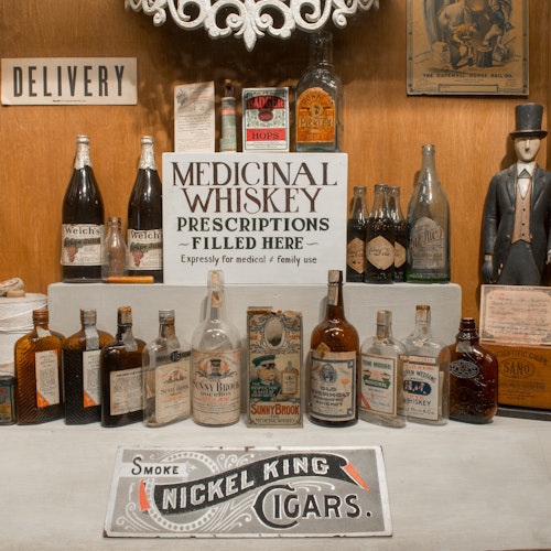 书架上摆放着各种威士忌瓶，挂着一个写着“药用威士忌”的牌子，还有一个写着“送货”的牌子。穿着西装和高顶礼帽的模特儿。
