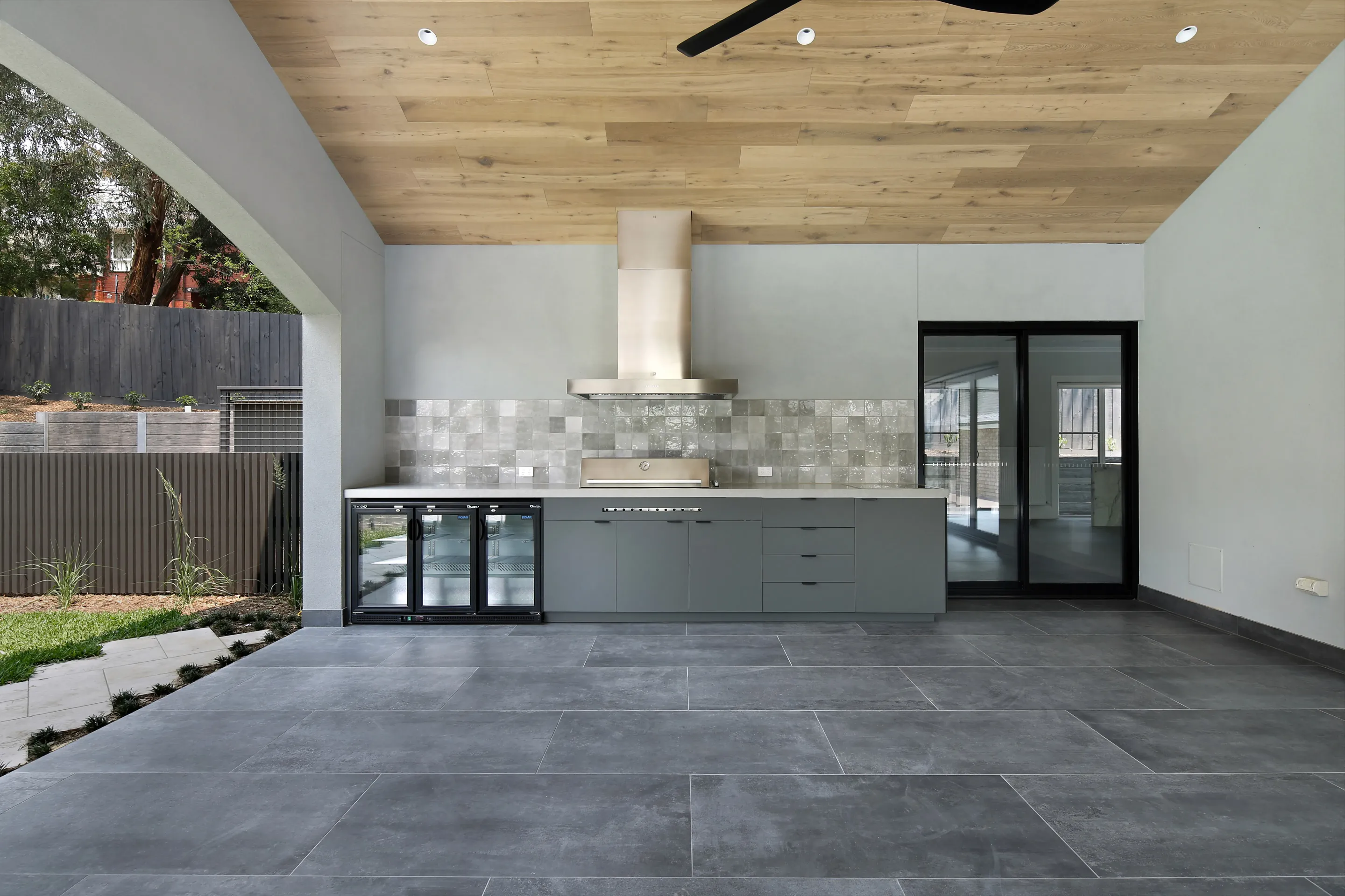 Outdoor kitchen with a timber-lined ceiling, stainless barbecue and rangehood, grey tile splashback, beverage fridges and a black sliding door, opening to a landscaped yard