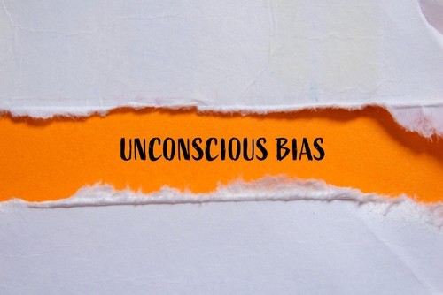 White paper torn open to reveal the words "UNCONSCIOUS BIAS" printed in bold black text on a bright orange background beneath.