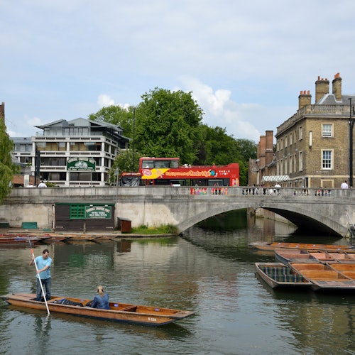 A person punts a boat on a river near a bridge with a red double-decker bus and buildings in the background.