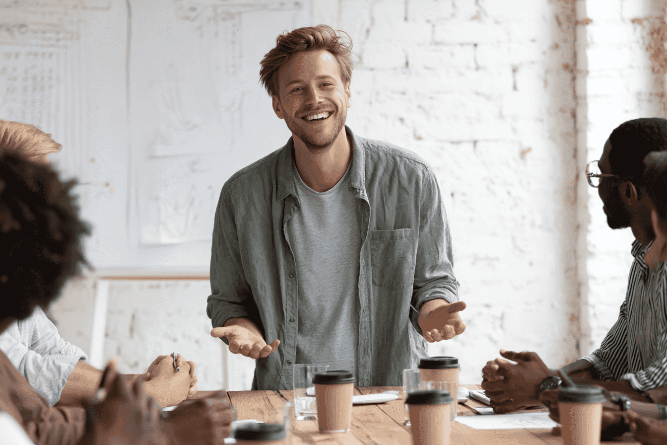 a young man is standing in front of his team, smiling and talking to them while sitting at the table with coffee cups on it.