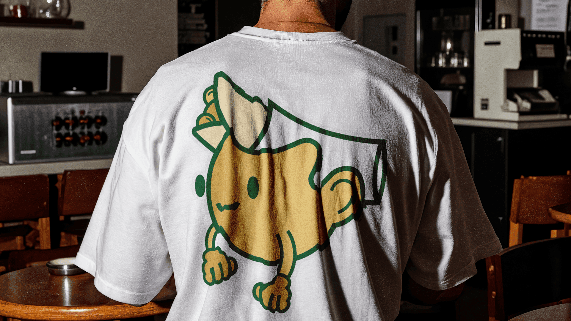 Person wearing a white T-shirt featuring a large Boccone Café mascot illustration on the back, photographed inside a cozy café setting.