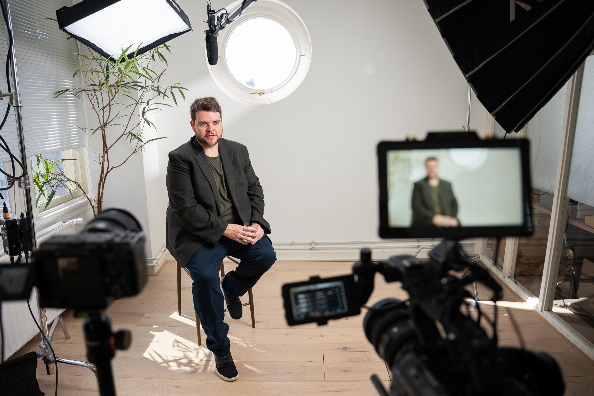 Behind-the-scenes wide shot of a video shoot showing a man in a dark blazer seated on a stool between two cameras with monitors, softbox lighting, and a round porthole window behind him.