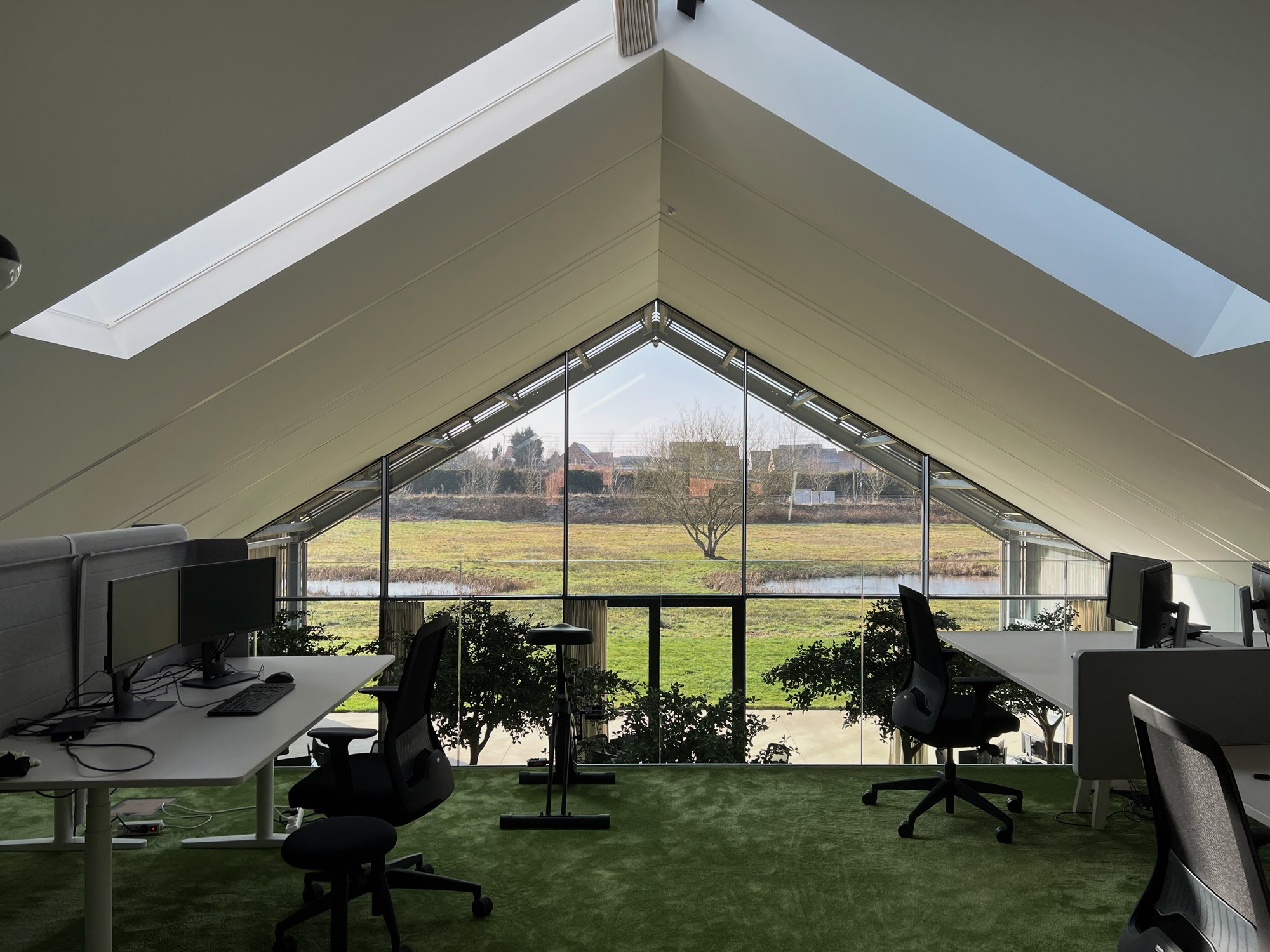 Het lichte interieur van een modern kantoor met een puntdak. Er staan witte zit-sta bureaus met dubbele monitoren op een groen tapijt. Door de grote glazen pui heb je een weids uitzicht over velden en een vijver.