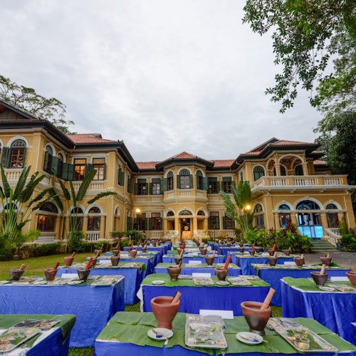 Deretan meja dengan penutup hijau serta lesung dan alu tanah liat disusun di hadapan bangunan besar gaya kolonial berwarna kuning.