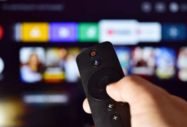 A hand holds a remote control pointed at a smart TV screen displaying various streaming app icons and options.