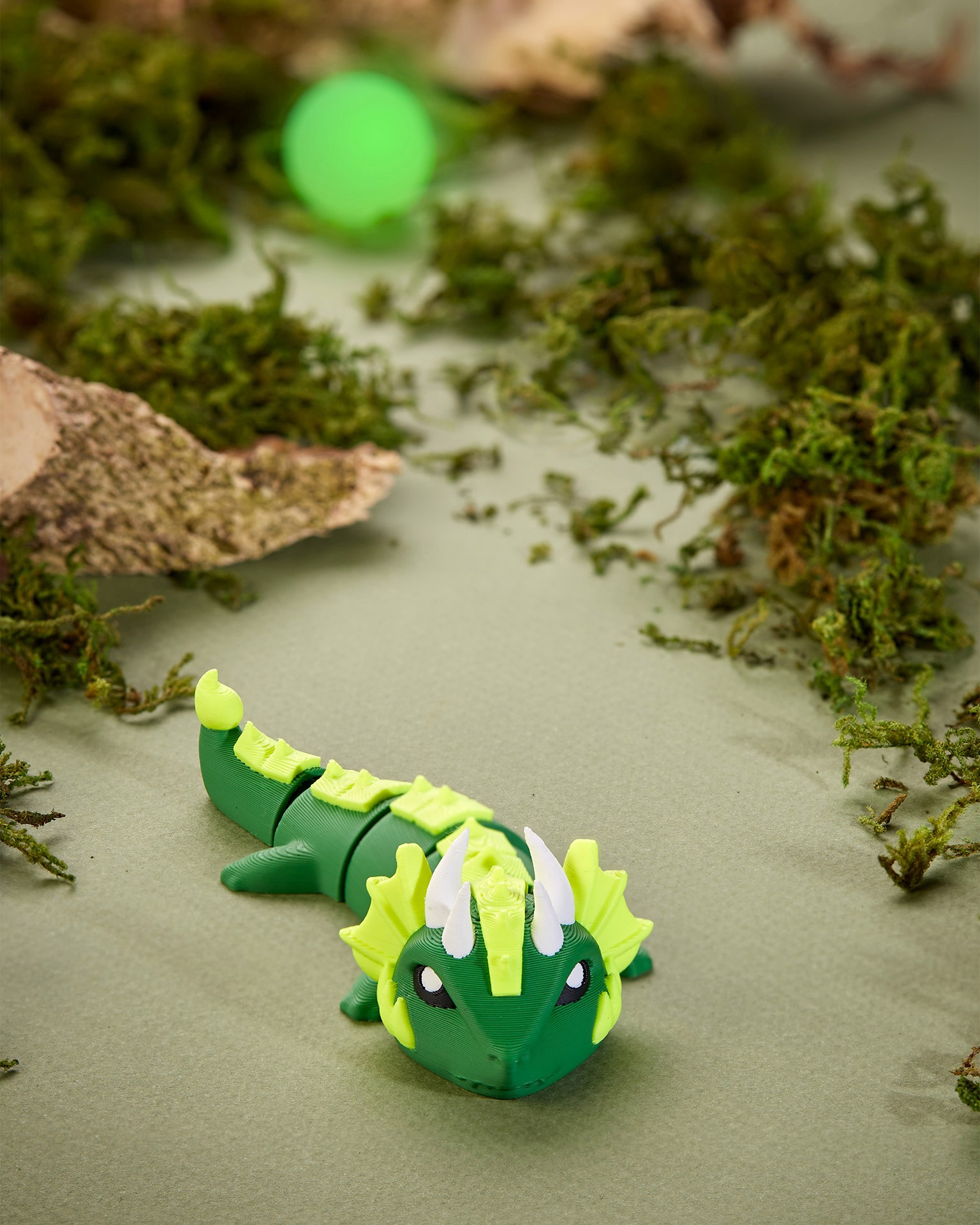 A small, green 3D-printed dragon figurine with intricate scales and horns is nestled among mossy branches on a muted green surface.